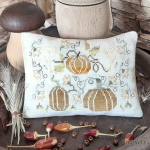 May include: A white pillow with a cross-stitch design featuring three pumpkins, vines, and leaves. The pillow is on a brown tray with dried flowers and wooden beads.