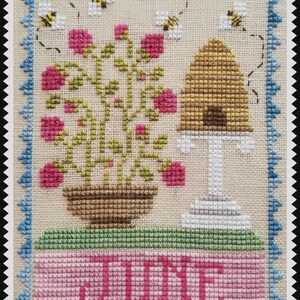 May include: A cross-stitch embroidery on beige fabric featuring a potted plant with pink flowers, a golden beehive on a white stand, and three bees. The word JUNE is stitched in pink at the bottom, all framed by a blue scalloped border.