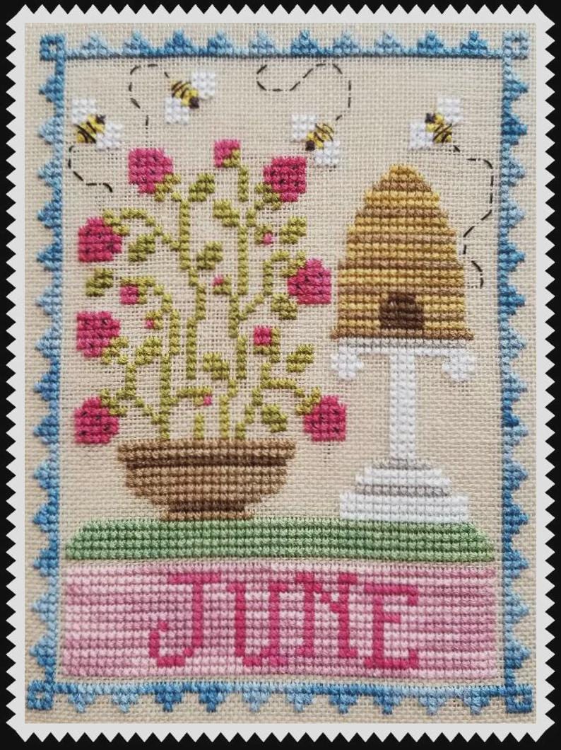 May include: A cross-stitch embroidery on beige fabric featuring a potted plant with pink flowers, a golden beehive on a white stand, and three bees. The word JUNE is stitched in pink at the bottom, all framed by a blue scalloped border.
