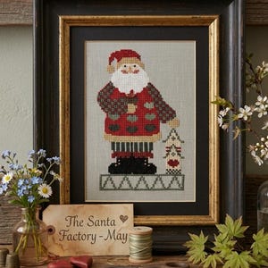 Può includere: Un ricamo a punto croce incorniciato di Babbo Natale con un cappotto a motivi rossi e verdi, che tiene in mano una piccola casa, in una cornice nera e oro. Un'etichetta di carta riporta la scritta The Santa Factory - May con un simbolo a cuore.