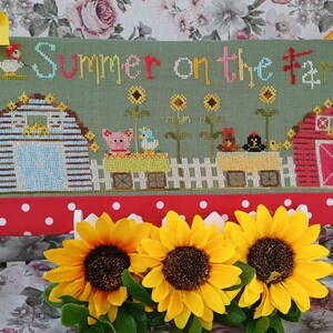 Op de afbeelding: Een rechthoekig kruissteek borduurwerk op saliegroene stof met de tekst Summer on the farm, twee schuren, zonnebloemen, een varken en vogels. Versierd met gele linten, lieveheersbeestje-bedels en een rode rand met witte stippen.