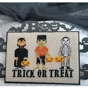 Może przedstawiać: Wyszywana krzyżykami dekoracja na Halloween przedstawiająca kota, Frankensteina i szkielet, każdy z dynią. Słowa "TRICK OR TREAT" są wyszyte poniżej. Dekoracja jest oprawiona w czarne wycinanki duch&oacute;w, nietoperzy i dyni.