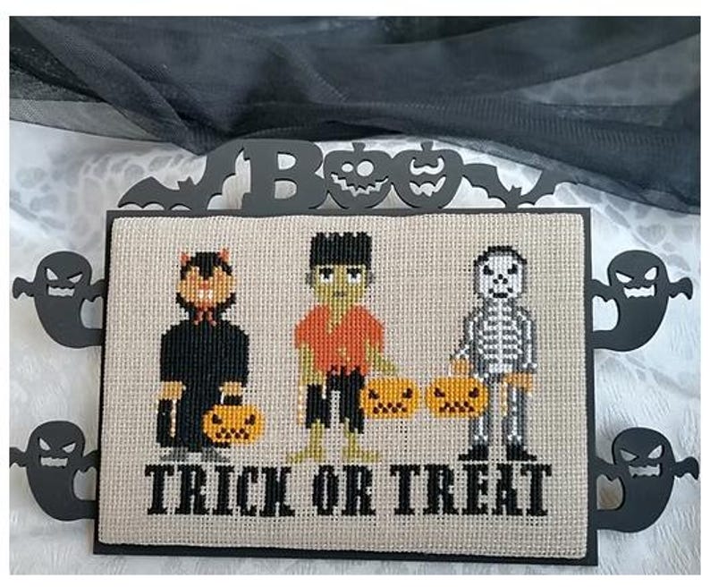 Może przedstawiać: Wyszywana krzyżykami dekoracja na Halloween przedstawiająca kota, Frankensteina i szkielet, każdy z dynią. Słowa "TRICK OR TREAT" są wyszyte poniżej. Dekoracja jest oprawiona w czarne wycinanki duch&oacute;w, nietoperzy i dyni.