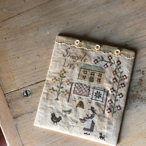 May include: A beige fabric pouch with a cross-stitched design featuring a house, flowers, and the words "Simple Life." The pouch is secured with three small buttons and has a decorative edge. A chicken, birds, and a dog are also depicted.