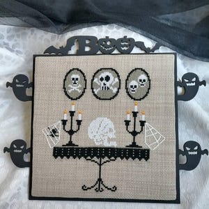 Może przedstawiać: Praca haftu krzyżykowego o tematyce Halloween z trzema wzorami czaszek i skrzyżowanych piszczeli. Grafika zawiera st&oacute;ł ze świecami, czaszką i pajęczynami. Czarne wycięcia duch&oacute;w obramowują grafikę, z napisem "Boo" na g&oacute;rze.