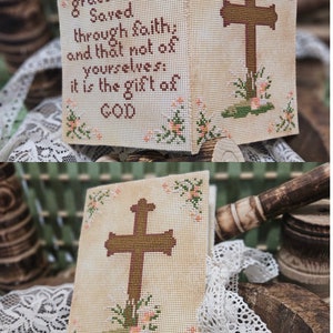 May include: A cross stitch embroidery design featuring a cross with a quote: "For by grace are ye Saved through faith; and that not of yourselves: it is the gift of GOD". The cross is brown and the background is beige with a floral border.