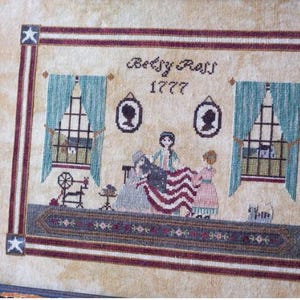May include: Framed cross-stitch depicting Betsy Ross and others creating the American flag. The scene includes windows with curtains, silhouettes, and the text "Betsy Ross 1777". The frame has red and white stripes and stars.