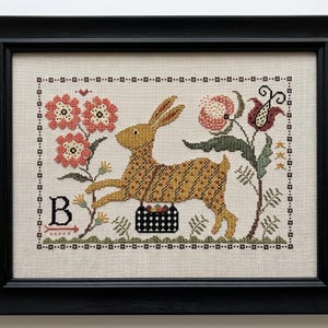 Könnte beinhalten: Eine gerahmte Kreuzstichstickerei mit einem goldbraunen Hasen, der über einen schwarzen Korb voller bunter Gegenstände springt. Das Design umfasst rosa und rote Blumen, grünes Blattwerk und den Buchstaben B mit einem kleinen roten Pfeil auf beigem Stoff.