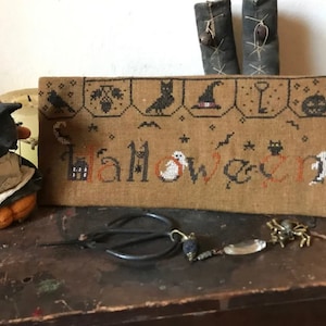 May include: A brown cross-stitch sampler with the word "Halloween" embroidered in orange and black thread. The sampler is surrounded by various Halloween-themed motifs, including bats, ghosts, and a witch's hat.