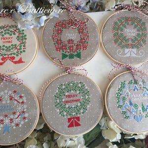 Op de afbeelding: Zes ronde houten borduurringen met kruissteekstof voorzien van kerstkransontwerpen. Elk stuk bevat de tekst Merry Xmas of Merry Christmas in rode of blauwe draad, geaccentueerd met rode, groene en witte decoratieve motieven.