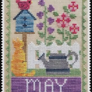 May include: A cross-stitch embroidery on beige fabric featuring a blue birdhouse with a pink bird, purple and pink flowers, a white picket fence, a grey watering can, and an orange cat. The word MAY is stitched in white on a purple banner.