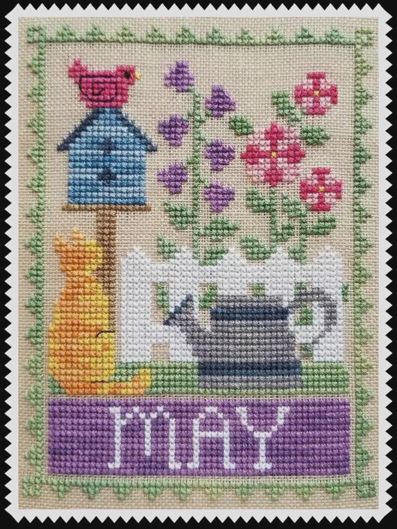 May include: A cross-stitch embroidery on beige fabric featuring a blue birdhouse with a pink bird, purple and pink flowers, a white picket fence, a grey watering can, and an orange cat. The word MAY is stitched in white on a purple banner.