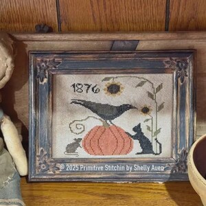 May include: A framed cross-stitch sampler featuring a black crow perched on an orange pumpkin, a black cat, a mouse, and a sunflower. The text 1876 and 2025 Primitive Stitchin by Shelly Auen are visible. The piece is in a dark, distressed wooden frame.