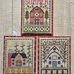May include: Three framed cross-stitch samplers depicting farm scenes. The top sampler says "SUMMER" above a red barn with sunflowers. The other samplers show barns with trees and flowers. Each sampler has a decorative border.