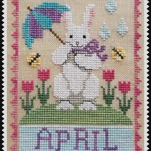 May include: A cross-stitch embroidery on beige fabric featuring a white rabbit holding a purple umbrella and a small purple object. The scene includes pink tulips, yellow bees, blue raindrops, and the word APRIL in purple, all framed by a pink border.