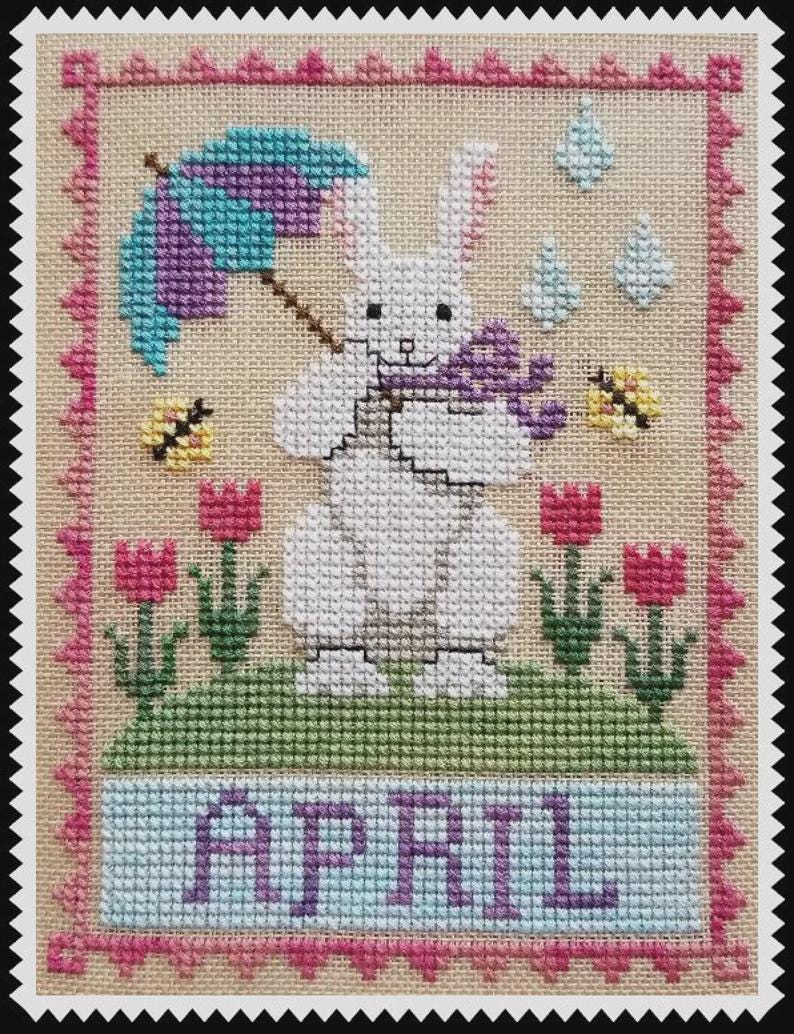 May include: A cross-stitch embroidery on beige fabric featuring a white rabbit holding a purple umbrella and a small purple object. The scene includes pink tulips, yellow bees, blue raindrops, and the word APRIL in purple, all framed by a pink border.