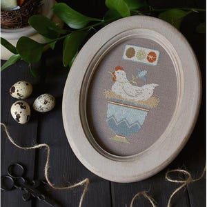 May include: An oval wooden frame containing a cross-stitch embroidery on grey fabric. The design features a white hen sitting in a blue patterned egg cup, a small blue bird, and three small coloured circles arranged in a horizontal row above the hen.