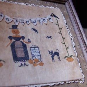 Op de afbeelding: Een ingelijste kruissteek met Halloween-thema met een heks, een zwarte kat en pompoenen. De banner luidt "HALLOWEEN". De heks houdt een drankfles vast bij een bord met "SPELL POTION". Het kunstwerk zit in een houten lijst.