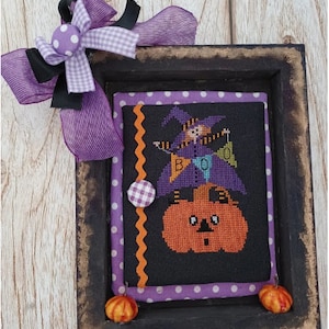 以下が含まれることがあります： 「BOO」の文字とカボチャに乗った魔女が描かれた、ハロウィーンのクロスステッチの額縁。 黒いフレームは、使い古したような仕上げで、紫色の水玉模様の縁取り、リボン、小さなカボチャの装飾が施されています。 季節感あふれるお祝いのデコレーションです。