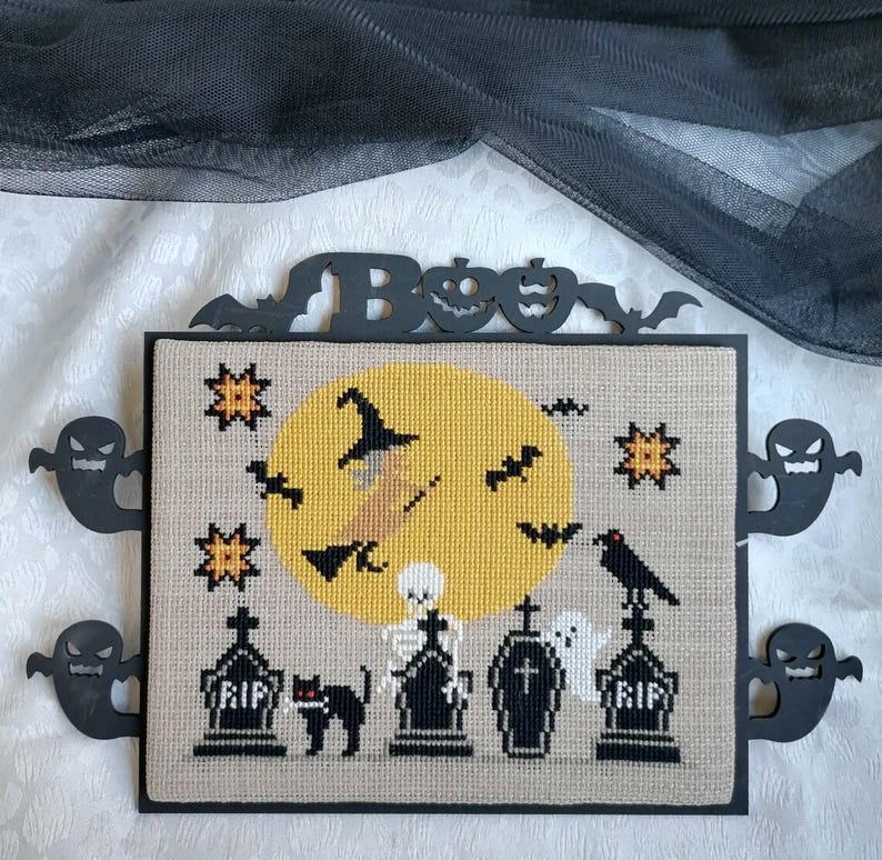 Może przedstawiać: Oprawiony haft krzyżykowy na Halloween przedstawiający wiedźmę na miotle przed ż&oacute;łtym księżycem, z nietoperzami, gwiazdami i czarnym kotem. Rama ma znak "Boo" i wycięcia duch&oacute;w. Haft krzyżykowy zawiera nagrobki i szkielet.