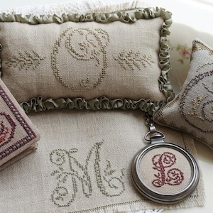 May include: A collection of embroidered items, including a square, a rectangular pillow, a small pillow, a fabric piece, and a round pendant. Each item features a different embroidered letter in shades of brown and red on a beige background.