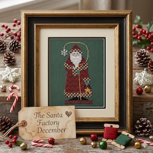 May include: A framed cross-stitch embroidery of Santa Claus in a red patterned coat on a dark green background. A tan paper tag in the foreground features the text "The Santa Factory - December" with a small heart icon printed on it.