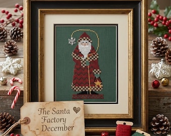 May include: A framed cross-stitch embroidery of Santa Claus in a red patterned coat on a dark green background. A tan paper tag in the foreground features the text "The Santa Factory - December" with a small heart icon printed on it.
