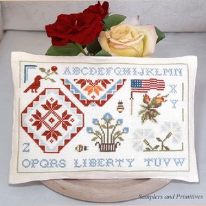 Könnte beinhalten: Ein rechteckiger, cremefarbener Kreuzstich-Sampler mit patriotischen und floralen Designs. Der Sampler zeigt eine amerikanische Flagge, Blumen und geometrische Muster in Rot, Blau und Orange. Das Wort "LIBERTY" ist unten eingestickt.