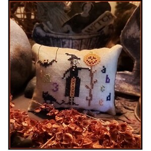 Pode incluir: Uma pequena almofada decorativa bege com um desenho de ponto cruz que apresenta uma bruxa, um gato preto, um morcego e uma abóbora. A bruxa está vestida com uma roupa preta e segura uma abóbora. A almofada tem o número "3" e as letras "a", "b" e "d".