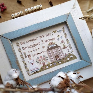 以下が含まれることがあります： 青と白のフレームに収められたクロスステッチ刺繍。刺繍には、家、女性、羊が描かれています。テキストには、「私の生活がシンプルになるほど、私は幸せになる」と書かれています。