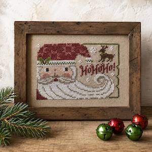 May include: A framed cross-stitch embroidery featuring a stylized Santa Claus with a red hat, white beard, and the text HOHOHO in red thread. A small brown reindeer is stitched above the text on a tan fabric background within a rustic wooden frame.