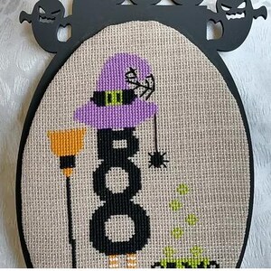 Może przedstawiać: Wz&oacute;r haftu krzyżykowego o tematyce Halloween, przedstawiający wiedźmę z fioletowym kapeluszem i słowem "BOO" w kolorze czarnym. Pomarańczowa miotła i pająk są r&oacute;wnież zawarte w projekcie. Projekt jest osadzony w owalnej ramie z wycięciami duch&oacute;w.
