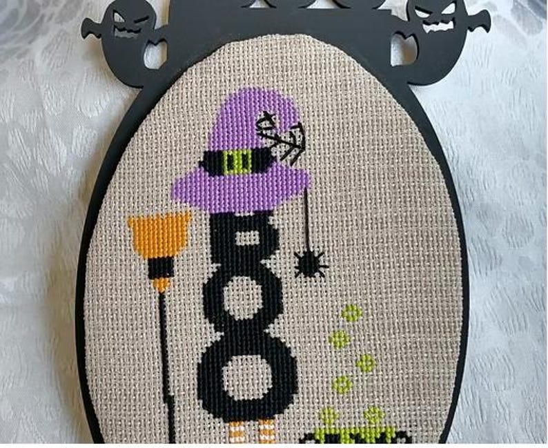 Może przedstawiać: Wz&oacute;r haftu krzyżykowego o tematyce Halloween, przedstawiający wiedźmę z fioletowym kapeluszem i słowem "BOO" w kolorze czarnym. Pomarańczowa miotła i pająk są r&oacute;wnież zawarte w projekcie. Projekt jest osadzony w owalnej ramie z wycięciami duch&oacute;w.