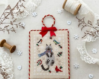 May include: A cross-stitch ornament depicting a grey cat wearing a blue scarf and a red bow. The cat is surrounded by snowflakes, holly, and birds. The ornament is framed with a red and gold trim.