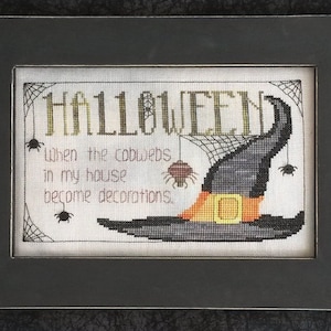 Op de afbeelding: Borduurwerk met kruissteek met de tekst "HALLOWEEN" en de zin "When the cobwebs in my house become decorations." Een zwarte heksenhoed met een oranje band en een spin die aan een web hangt, is afgebeeld.