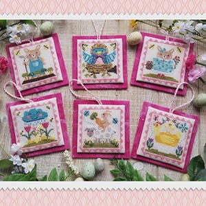 May include: Six square cross-stitch ornaments featuring embroidered spring motifs including a rabbit, a blue bird in a nest, a floral bunny, a rain cloud over tulips, a sheep with a carrot, and a yellow chick, each mounted on a bright pink felt backing.