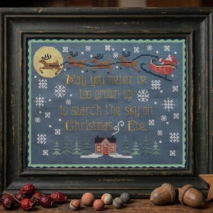 Peut inclure: Une broderie au point de croix encadrée sur tissu bleu foncé représentant une lune jaune, un traîneau rouge avec des rennes et le texte : May you never be too grown up to search the sky on Christmas Eve. Le motif inclut des flocons blancs.