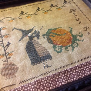 Peut inclure: Broderie au point de croix représentant une sorcière, une citrouille et l'inscription "Boo! Boo!". La sorcière porte un chapeau et une robe noirs, près d'une grosse citrouille orange. Un arbre avec des citrouilles et des oiseaux est à gauche. L'œuvre est encadrée.