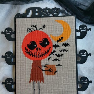 Może przedstawiać: Haft krzyżykowy z motywem Halloween przedstawiający uśmiechniętą postać z dyniową głową trzymającą torbę na smakołyki, z p&oacute;łksiężycem i nietoperzami. Grafika jest oprawiona w czarne akcenty i wycięcia duch&oacute;w, ze słowem "Boo" na g&oacute;rze.