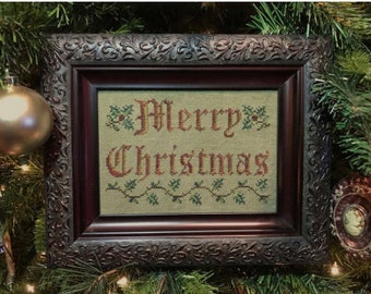 Pode incluir: Obra de arte bordada em ponto cruz com as palavras "Merry Christmas" em letras vermelhas. A obra está emoldurada em uma moldura marrom escura e ornamentada. A vegetação e um enfeite dourado são visíveis no fundo, sugerindo um tema de feriado.