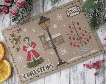 Pode incluir: Design de ponto cruz representando uma cena de Natal com uma mulher de casaco vermelho, um lampião, um edifício e uma árvore com bagas vermelhas. A palavra "CHRISTMAS" é costurada na parte inferior e o ano "1863" é exibido.