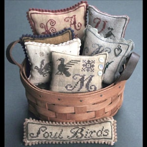 Peut inclure: Un panier rond en bois rempli de plusieurs petits coussins carrés en tissu beige ornés de lettres, d'oiseaux et de motifs brodés au point de croix. Une pièce rectangulaire en tissu avec le texte "Soul Birds" est placée devant le panier.