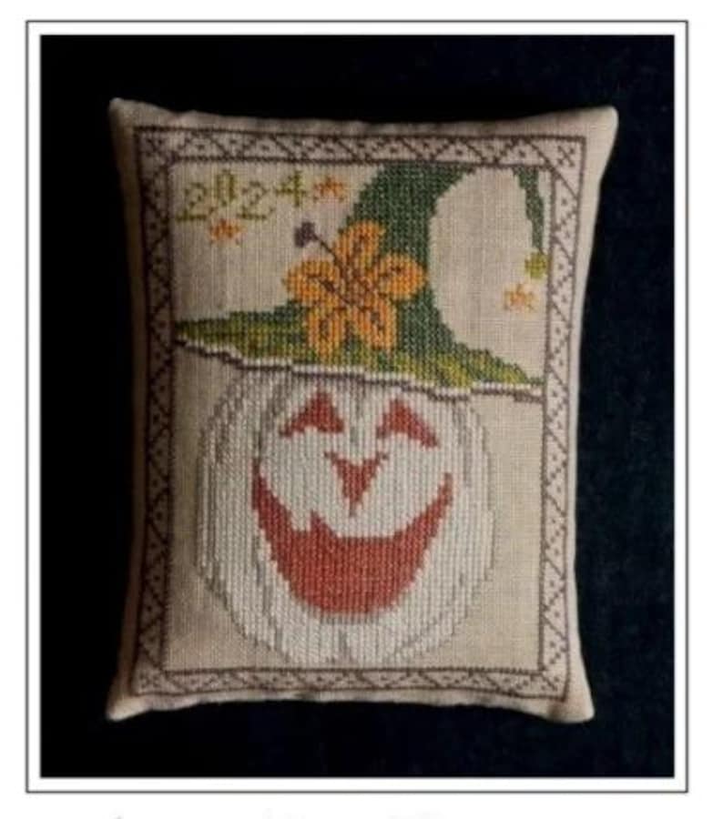 May include: A decorative, rectangular pillow with a cross-stitch design featuring a smiling pumpkin wearing a green witch hat. The pumpkin has a red-orange mouth and is framed by a decorative border. The text "2024" is stitched above the hat.