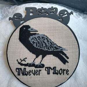 Może przedstawiać: Okrągła, haftowana krzyżykami dekoracja na Halloween z czarnym krukiem z szarymi detalami i napisem "Nevermore". Czarna, obramowana dekoracja ma czarne wycięcie ze słowem "Boo" oraz kształtami duch&oacute;w i dyni.