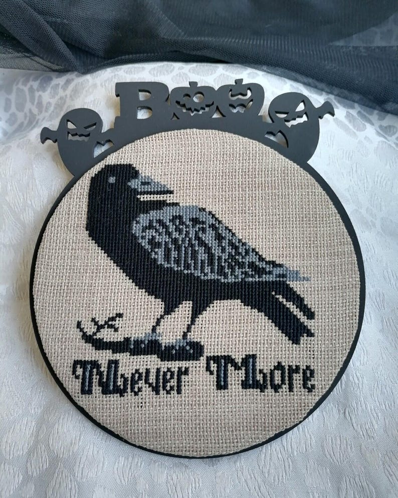 Może przedstawiać: Okrągła, haftowana krzyżykami dekoracja na Halloween z czarnym krukiem z szarymi detalami i napisem "Nevermore". Czarna, obramowana dekoracja ma czarne wycięcie ze słowem "Boo" oraz kształtami duch&oacute;w i dyni.