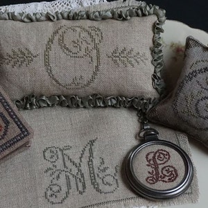 May include: A set of four embroidered pillows with different designs. Each pillow features a different letter of the alphabet, with a floral design around the letter. The pillows are made of a light brown fabric and have a rustic look.