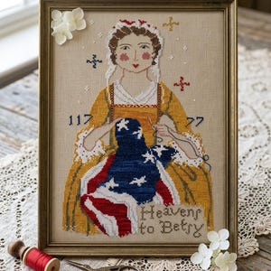 May include: Framed cross-stitch artwork featuring a woman in a yellow dress stitching an American flag. The piece includes the text "Heavens to Betry" and the numbers "117" and "77". The frame is gold-colored.