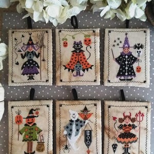 May include: A collection of six Halloween-themed cross-stitch designs. Each design features a different character, including witches, a black cat, and a pumpkin-headed figure. The designs are framed with a decorative stitch and have a black ribbon for hanging.