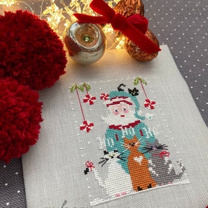Può includere: Un ricamo a punto croce raffigurante Babbo Natale con tre gatti bianchi, arancioni e grigi. Il disegno è su tessuto grigio chiaro, con la parola "myfanny" in corsivo. Elementi decorativi: un pompon rosso, ornamenti e luci.