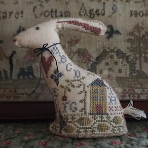 May include: A beige and brown embroidered rabbit with a black ribbon around its neck. The rabbit is sitting on a brown surface and has a small house embroidered on its side. The house has a blue roof and yellow windows.